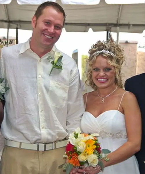 Bride and groom smiling, bride holding a bouquet of colorful flowers under a white tent.