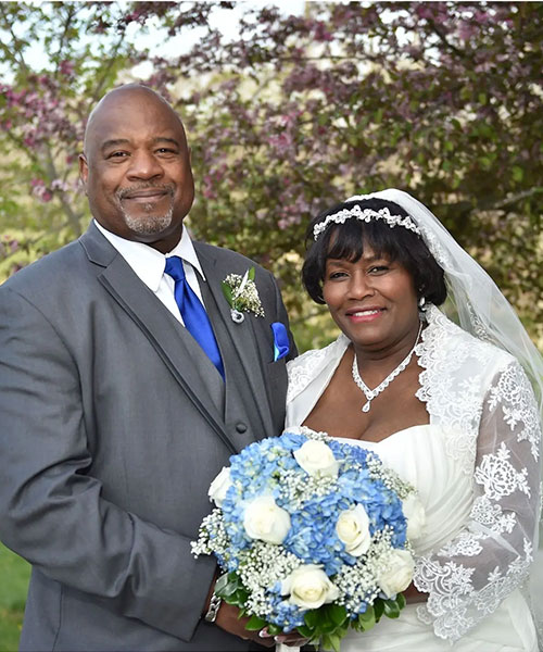 A couple dressed in wedding attire stands outdoors; the groom in a gray suit and the bride in a white dress holding flowers.