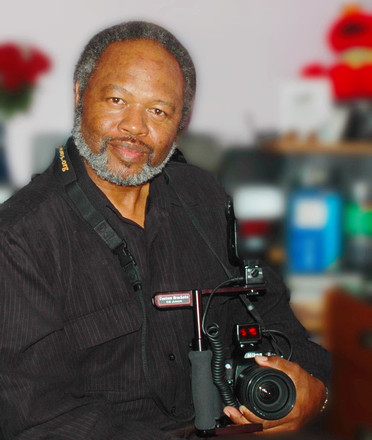 Man seated, holding a camera with a shoulder rig, and wearing a dark shirt.