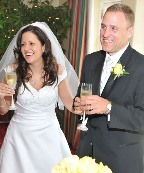 A bride and groom stand side by side, smiling and holding glasses of champagne. The bride wears a veil and white dress.